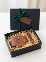 A brown leather motorcycle boot protector presented in an open black gift box filled with shredded paper, with the matching closed lid behind it and a green ribbon and tag