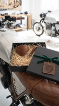 A motorcycle gift box set with leaft boot cover and keichain, displayed on a brown leather motorcycle seat in a workshop.