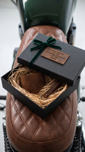 Close-up of brown leather motorcycle shifter boot protector and keichain, A black gift box with a dark green ribbon sits open on a brown quilted motorcycle seat. 