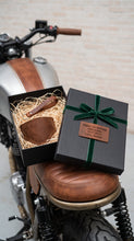 A black gift box with a green ribbon and a personalized motorcycle brown leather boot protector cover and leather keychain