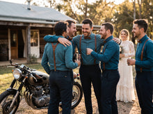 A group of five men celebrating outdoors in front of a rustic cabin, with a motorcycle in the foreground and a bride in a white wedding gown in the background. The men are wearing matching teal shirts, dark trousers, brown suspenders, and brown leather bow ties.