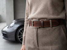 A close-up of a person wearing a brown leather belt with a silver buckle over a beige knit sweater and matching trousers, with a blurred gray sports car in the background.