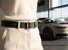 A close-up of a person wearing a black textured leather belt with a silver buckle, paired with cream-colored trousers. The background shows a blurred Porsche inside a modern car showroom.