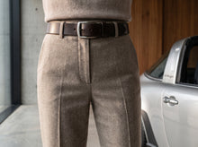 A close-up of a person wearing high-waisted taupe wool trousers and a dark brown leather belt, standing next to a white car in an indoor setting.