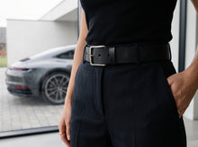 A close-up of a person wearing a black leather belt with a silver-toned rectangular buckle, paired with dark trousers and a black top. The background shows a blurred gray sports car outside a modern building.