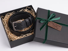 A black leather belt with a silver buckle presented in an open black gift box filled with shredded paper, alongside a closed black gift box tied with a green ribbon and a wooden tag that reads 'Merry Christmas, Daughter, Love Dad'.