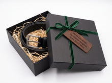 A black leather belt with a silver buckle presented in an open black gift box with shredded paper filler. The closed lid is tied with a dark green ribbon and has a brown engraved tag that reads, 'Merry Christmas, my dearest Daughter, Love, Dad & Mom'.