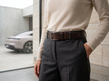 A close-up lifestyle shot of a person wearing a dark brown leather belt with a silver-toned rectangular buckle, styled with a cream knit sweater and dark gray trousers.