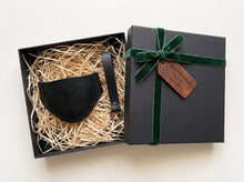A black leather motorcycle shifter boot protector with keichain presented in an open black gift box filled with shredded paper, next to a closed black gift box tied with a green ribbon and a brown tag that reads 'Happy Anniversary, My love, Your wife'.