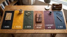 Six leather desk mats in different colors are arranged on a wooden table, each labeled with a color name and styled with office accessories like a notebook, glasses, and a pen. The mats are labeled: 1.