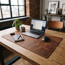 A large brown leather desk mat with 'A.M Creative Studio' embossed in the corner, styled on a wooden table with a laptop, notebook, pen, mug, and plant in a bright office setting.