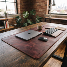 A large burgundy wine leather desk mat with 'ADAM C. CROTTY' printed in the corner, styled on a rustic wooden desk with a laptop, keyboard, mouse, and coffee cup in a modern loft office setting.
