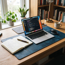 A styled home office desk setup featuring a large dark blue leather desk mat with a silver laptop, an open notebook with a pen, a computer mouse, and a mug, set in a warm, sunlit room with a window, plants, and bookshelves.