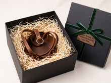 Close-up of leather plant hanger straps and metal ring, full grain leather texture inside an open black gift box filled with shredded paper, with a closed black gift box and green ribbon beside it on a light background.