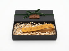 A personalized leather keychain with an engraved name and phone number, presented inside an open black gift box with a green ribbon and shredded paper.