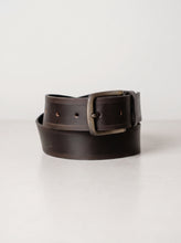 A dark brown leather belt with a distressed finish and a square bronze-toned metal buckle, coiled on a light gray surface.