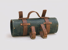 A dark green canvas cylindrical tool roll with brown leather straps, handles, and buckles, shown on a light gray background.