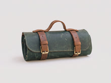 A dark green rolled leather tool roll with brown leather straps and handles, featuring brass buckles, shown on a light gray background.