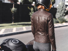 A women viewed from behind wearing a brown quilted leather jacket, gray jeans, and a black helmet while standing outdoors next to a motorcycle.