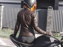 A women wearing a brown quilted leather jacket and a black helmet sits on a black motorcycle, photographed from behind in an outdoor setting with a fence in the background.