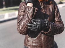 A person wearing a brown leather jacket and black leather gloves, standing outdoors on a street with blurred background.