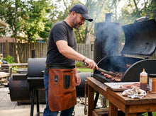 A man wearing a dark gray t-shirt, blue jeans, and a brown leather apron stands outdoors next to a smoking charcoal grill, using tongs to flip meat. A wooden table with cooking ingredients and towels is in the foreground, with another grill and trees in the background.