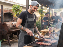 A man wearing a gray waxed canvas leather apron and a gray baseball cap grills ribs outdoors at a backyard barbecue with friends. A smoker and string lights are visible in the background.