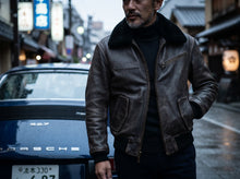 A man wearing a aviator brown leather jacket with a black shearling-style collar over a black turtleneck, standing on a wet city street in front of a blue Porsche.