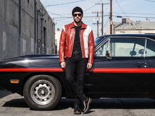 A man wearing a red and white leather jacket, black beanie, sunglasses, and black pants stands next to a black classic muscle car with a red stripe, in an outdoor urban setting.
