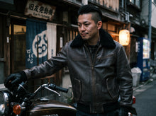 A man wearing a dark brown leather motorcycle aviator jacket with a black shearling-style collar stands next to a motorcycle in a narrow Japanese street.