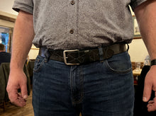 A close-up of a man wearing a dark brown leather belt with a geometric diamond pattern and a silver buckle, paired with blue jeans and a patterned short-sleeve button-up shirt.