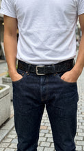 A man wearing a white t-shirt and dark blue jeans, featuring a black belt with a geometric diamond pattern and a silver buckle.