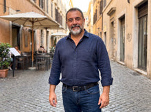 A man with a gray beard and mustache wearing a navy blue button-down shirt and dark blue jeans, standing in a cobblestone street with warm-toned buildings and cafe umbrellas in the background.