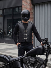 A person wearing a black motorcycle vest over a black long-sleeve shirt and black helmet stands beside a black cruiser motorcycle on a city sidewalk in front of a brick and metal building.