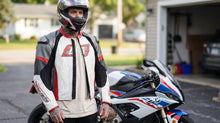 A man wearing a full-face helmet and a white, red, and black Leatt motorcycle riding jacket stands in a residential driveway next to a white, blue, and red BMW sport motorcycle.