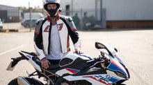 A man in a white, black, and red BMW racing leather jacket and full-face helmet stands on a paved road next to a white, blue, and red BMW S1000 sport motorcycle.