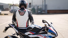A motorcyclist wearing a white, black, and red Leatt helmet and riding suit sits on a BMW sport motorcycle in an outdoor paved area.