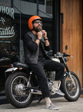 A man wearing a black leather vest, black shirt, black pants, and a bright orange helmet sits on a black cruiser motorcycle in front of a storefront with wood paneling.