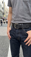 A close-up lifestyle shot of a man wearing a black belt with a geometric pattern and a silver buckle, paired with dark blue jeans and a gray t-shirt, standing on a city street.