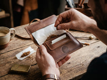 open brown leather tobacco pouch on a wooden table, with loose rolling tobacco, rolling papers, a lighter, and coffee nearby.