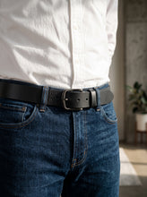 A close-up of a person wearing a black leather belt with a silver-toned rectangular buckle, tucked into dark blue jeans and a white button-up shirt.