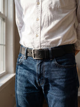 A close-up of a person wearing a black leather belt with a silver-toned rectangular buckle, styled with dark blue jeans and a white button-up shirt in a sunlit indoor setting.