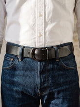 A close-up of a person wearing a black  leather belt with a silver-toned metal buckle, paired with dark blue jeans and a white button-down shirt.