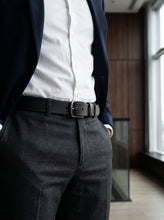 A close-up of a person wearing a black leather belt with a silver buckle, styled with a white dress shirt, navy blazer, and dark gray trousers in a modern indoor setting.