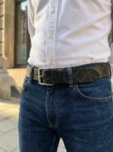 A close-up of a person wearing a black leather belt with a geometric cutout pattern and a silver buckle, styled with a white button-down shirt and blue jeans.