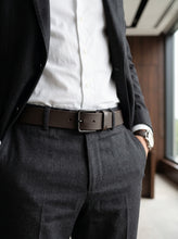 A close-up of a man wearing a dark brown leather belt with a silver-toned metal buckle, styled with a white dress shirt, dark gray suit jacket, and matching dark gray trousers.