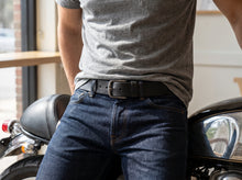 A close-up lifestyle shot of a man wearing a black leather belt with a silver buckle, styled with a gray t-shirt and dark blue jeans while leaning against a black motorcycle.