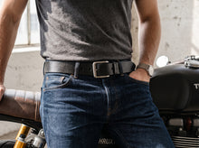 A man wearing a black leather belt with a silver-toned rectangular buckle, styled with a dark gray t-shirt and blue jeans, leaning against a motorcycle in a rustic indoor setting.