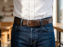 A close-up shot of a person wearing a dark brown leather belt with a silver-toned rectangular buckle, styled with a white button-down shirt and dark blue jeans in a softly blurred indoor setting.