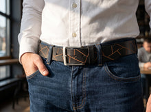 A close-up of a man wearing a black leather belt with a geometric copper-orange line pattern, styled with a white button-down shirt and blue jeans.
